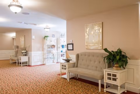 A well-lit hallway in an assisted living facility featuring a patterned carpet, beige walls with white wainscoting, a tufted beige bench with side tables on either side, one holding a potted plant and the other a decorative sailboat. In the background, there is a white bookshelf with various items and a framed picture on the wall.