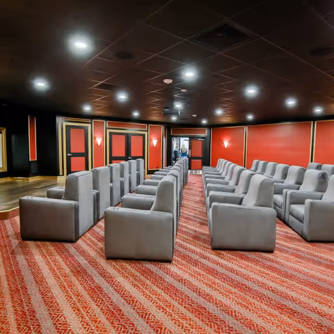 Interior view of a theater room with multiple rows of gray cushioned seats arranged on a red patterned carpet. The walls are painted red with black and gold trim, and the ceiling is dark with recessed lighting. A doorway is visible at the back of the room.
