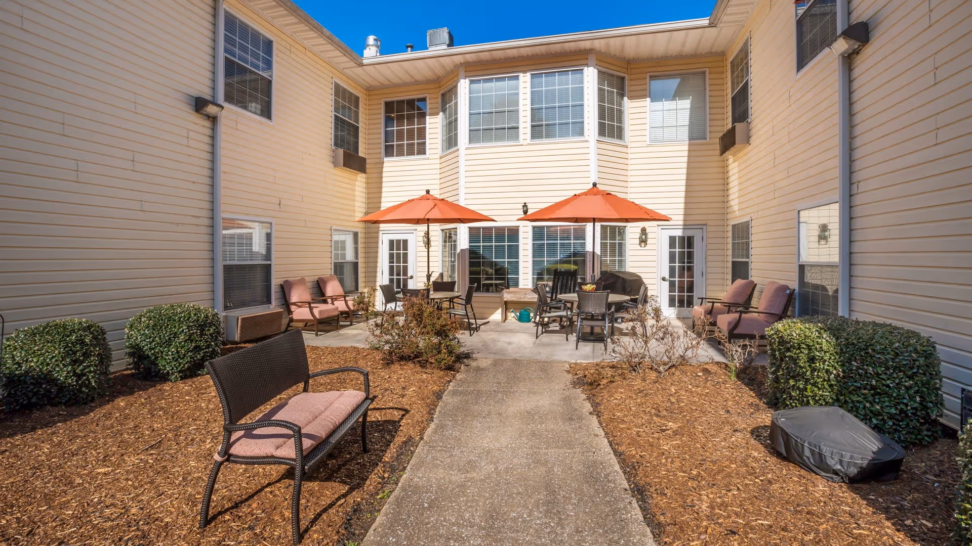 Outdoor courtyard area of a senior living facility with beige siding walls, several windows, and two orange patio umbrellas shading tables and chairs. There are cushioned chairs and benches arranged around the concrete patio, with mulch and trimmed bushes lining the walkway leading to the seating area.
