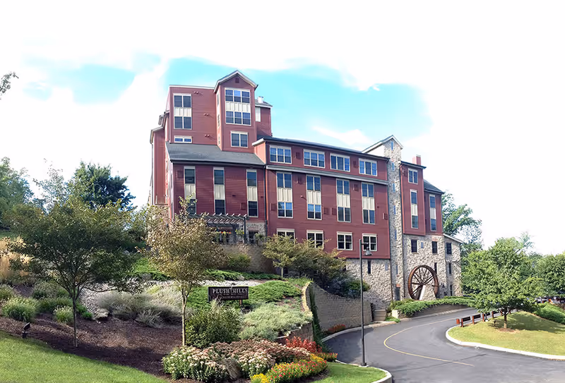 Red multi-story senior living building with stone accents, a decorative water wheel, landscaped gardens and a curved driveway.