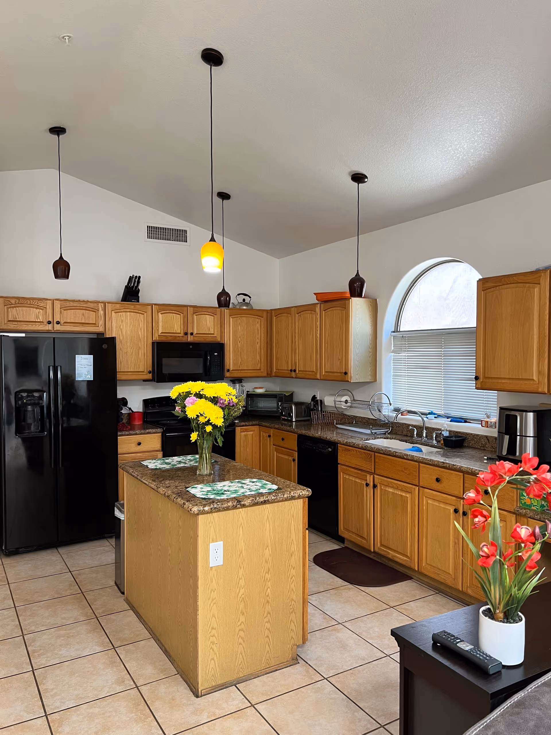 A kitchen with wooden cabinets, a black refrigerator, black microwave, and black dishwasher. There is a kitchen island with a vase of yellow flowers and two placemats. Three pendant lights hang from the ceiling, one with a yellow shade. A window with an arched top and closed blinds is above the sink. A small table with a remote control and a vase of red flowers is visible in the foreground.