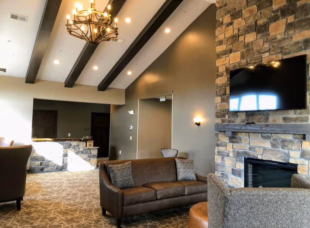 A cozy living room area in a retirement facility featuring a stone fireplace with a mounted flat-screen TV above it. The room has a high ceiling with exposed dark wooden beams and a decorative chandelier. There is a brown leather sofa with patterned cushions, a gray upholstered chair, and a stone half-wall in the background near a doorway.