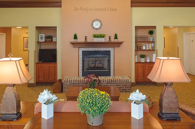 A cozy senior living common area with a central fireplace surrounded by built-in wooden shelves filled with books and decorative items. Two table lamps with patterned bases flank a wooden table adorned with potted plants and flowers. Above the fireplace is a clock and the phrase 'Our Family is Committed to Yours' on the wall.