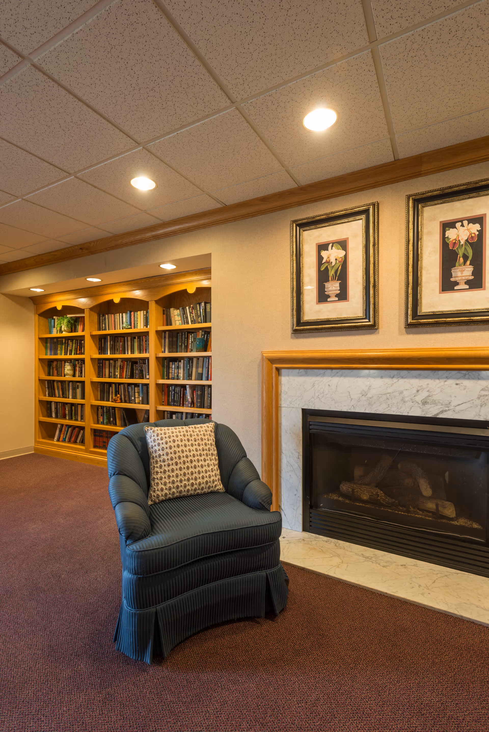 A cozy interior room featuring a blue upholstered armchair with a patterned cushion in front of a marble-framed fireplace. Behind the chair, there is a built-in wooden bookshelf filled with books, and two framed floral paintings hang above the fireplace. The room has a carpeted floor and recessed ceiling lights.