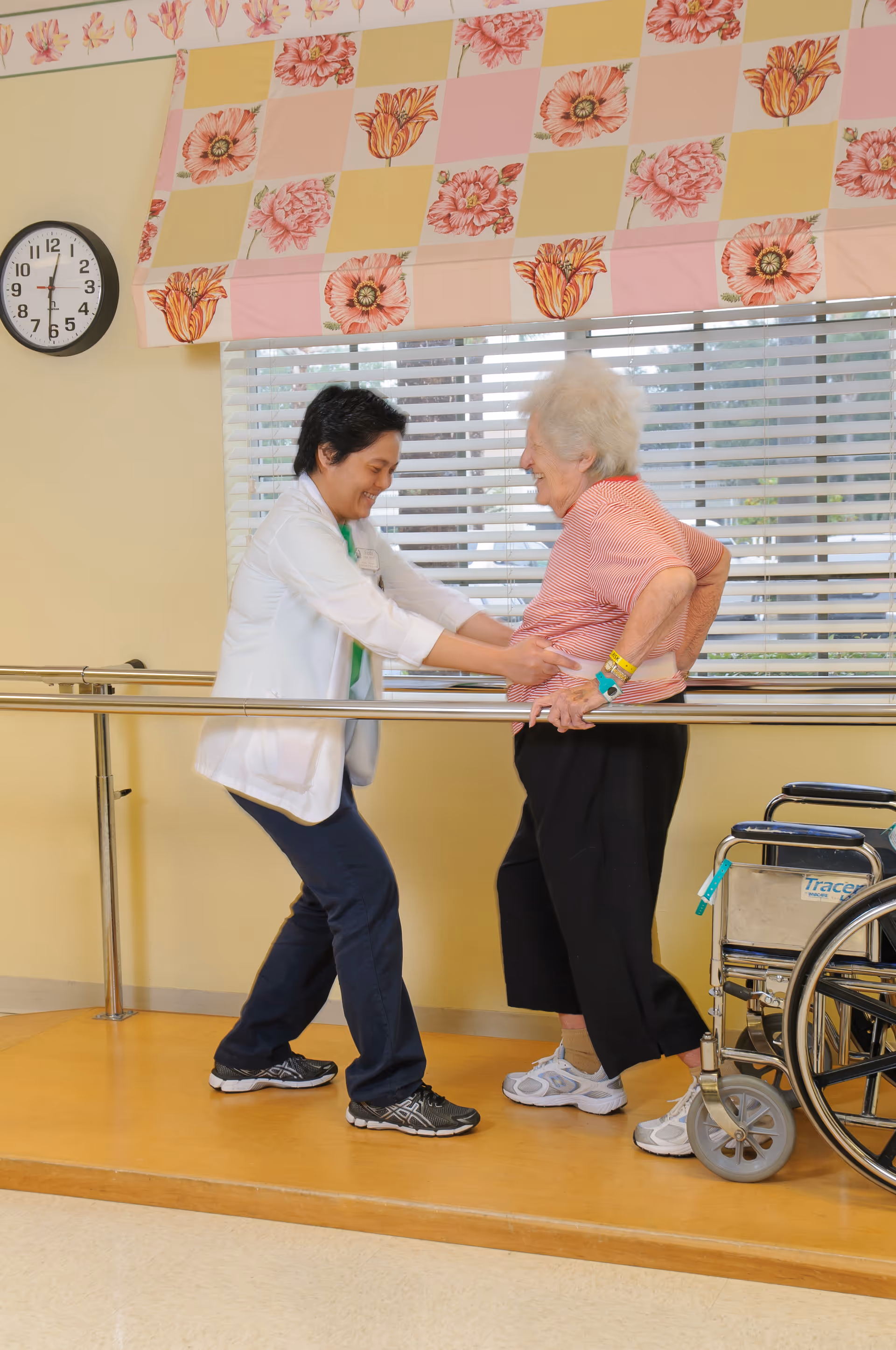 A healthcare worker assists an elderly woman with walking exercises using parallel bars in a rehabilitation or therapy room. The elderly woman is wearing a red striped shirt and black pants, and there is a wheelchair nearby. The room has a clock on the wall and floral patterned window valances.