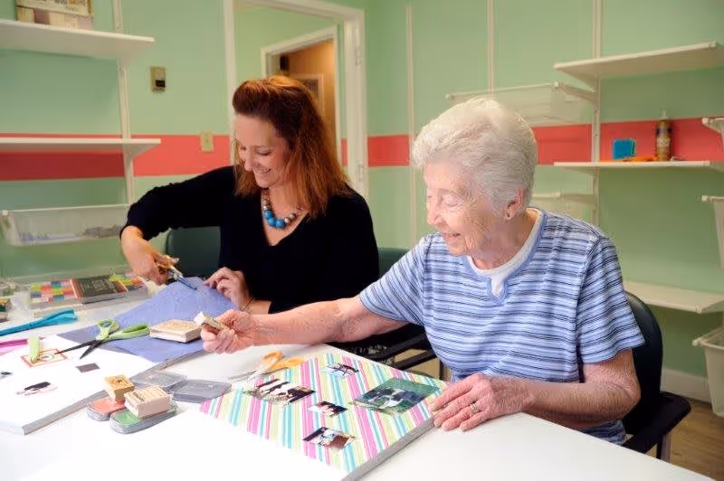 An elderly woman and a younger woman sitting at a table engaged in a crafting activity. The elderly woman is holding a stamp and working on a colorful striped scrapbook page with photos, while the younger woman is cutting fabric with scissors. They are in a room with light green walls and white shelves.