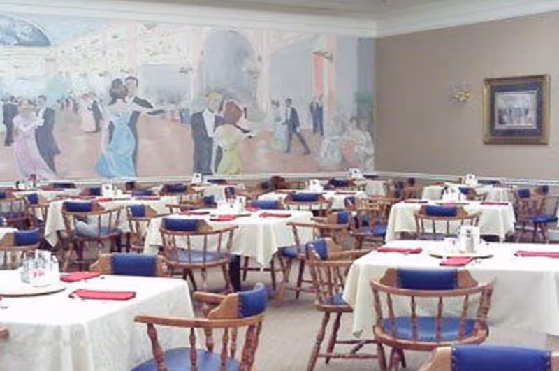 Dining room with multiple round tables covered with white tablecloths, each set with red napkins and condiments. Wooden chairs with blue cushions surround the tables. A large mural depicting people dancing in formal attire decorates one wall, and a framed picture hangs on another wall.