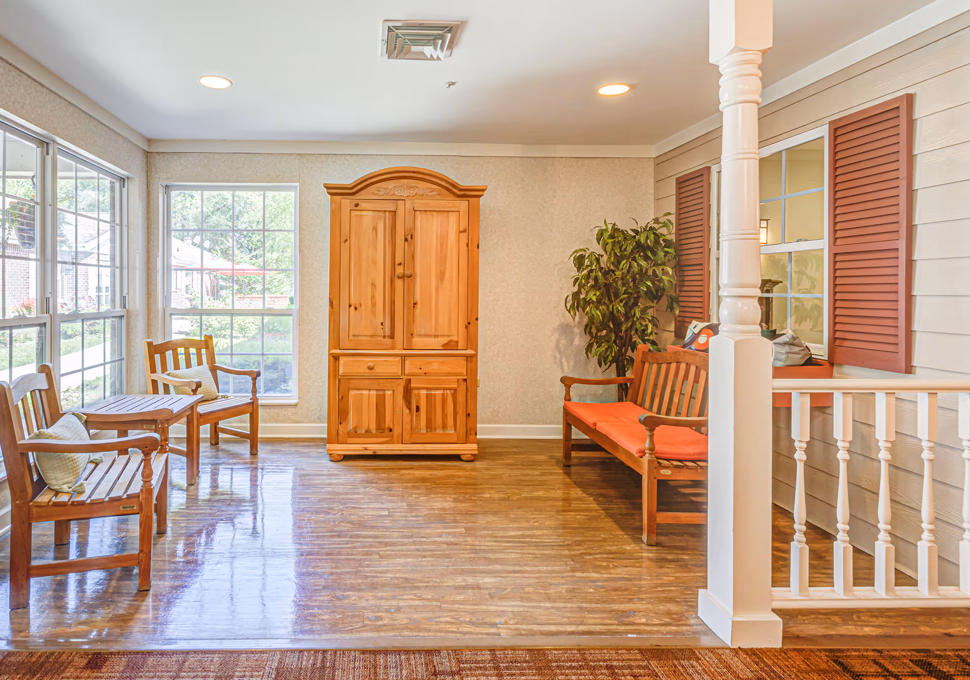 Sunlit interior seating area with wooden benches, a wooden armoire, a potted plant and large windows.