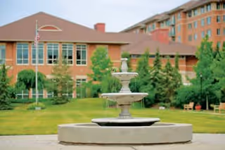 A multi-tiered water fountain in the foreground with a well-maintained lawn, trees, and shrubs. In the background, there is a large building with multiple windows and a brown roof, likely part of a senior living community.