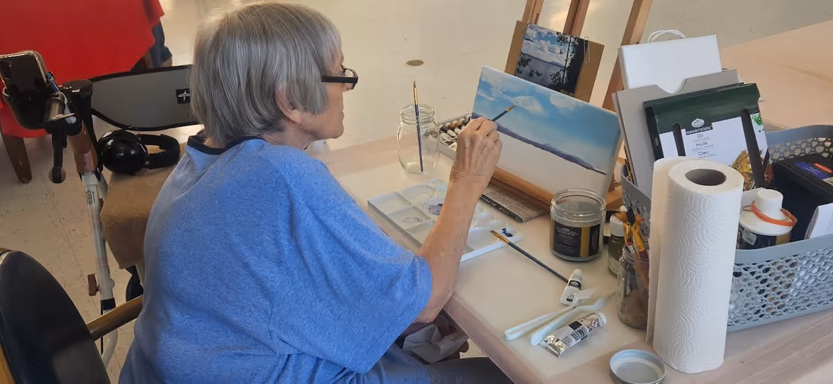 An elderly woman with short gray hair and glasses is seated at a table painting a landscape on a canvas. The table holds various painting supplies including brushes, paint tubes, a jar of water, paper towels, and a container with other art materials. The setting appears to be a well-lit indoor space.