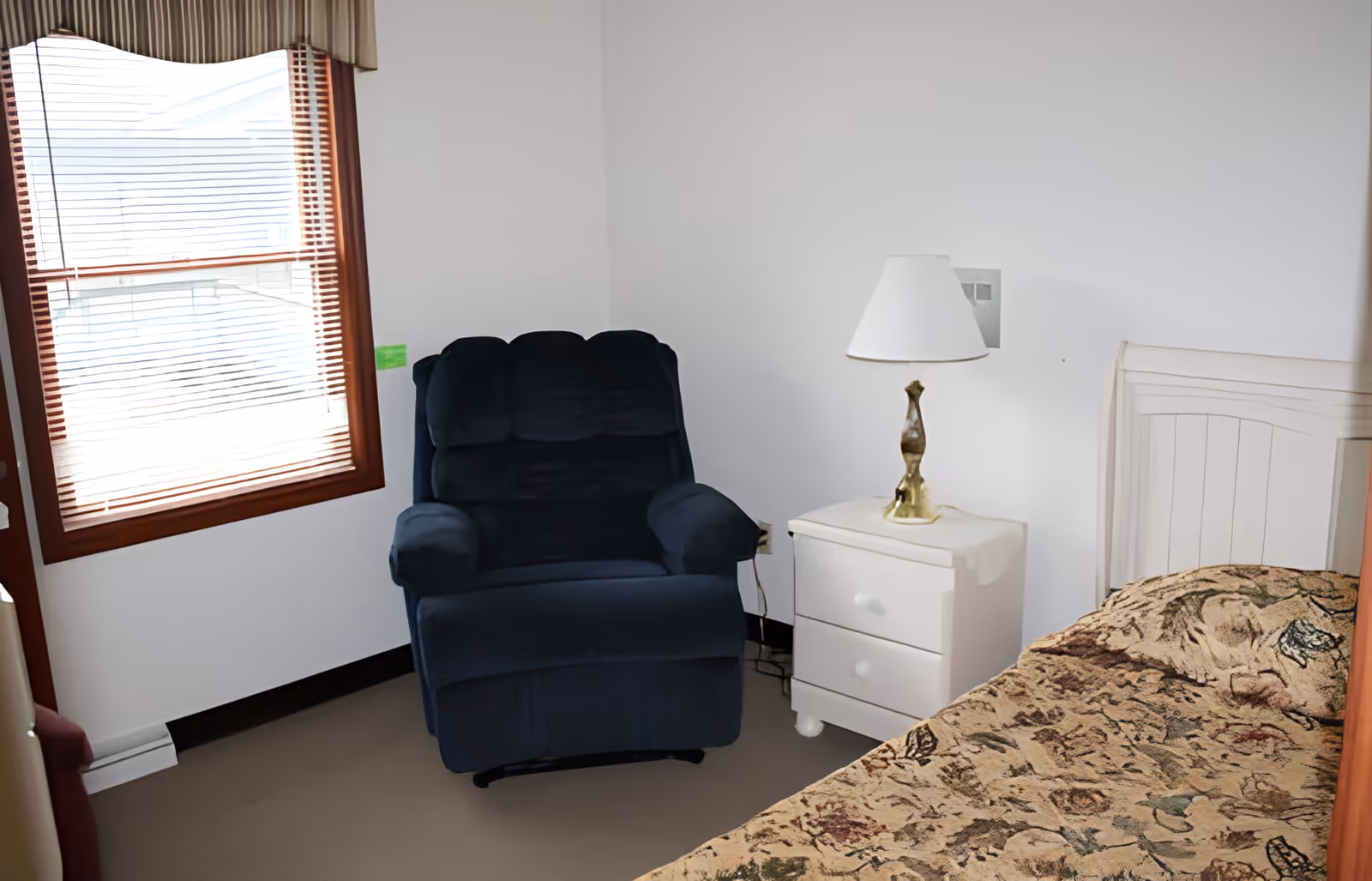 A small bedroom with a window covered by blinds and a valance, a dark blue upholstered armchair, a white nightstand with two drawers, a brass table lamp with a white lampshade, and a bed with a floral patterned bedspread.