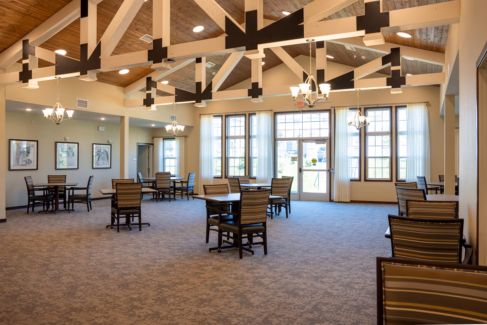 Spacious dining room in Boden Senior Living with multiple tables and chairs arranged on carpeted floor, large windows with sheer curtains allowing natural light, and a high wooden ceiling with exposed beams and hanging light fixtures.