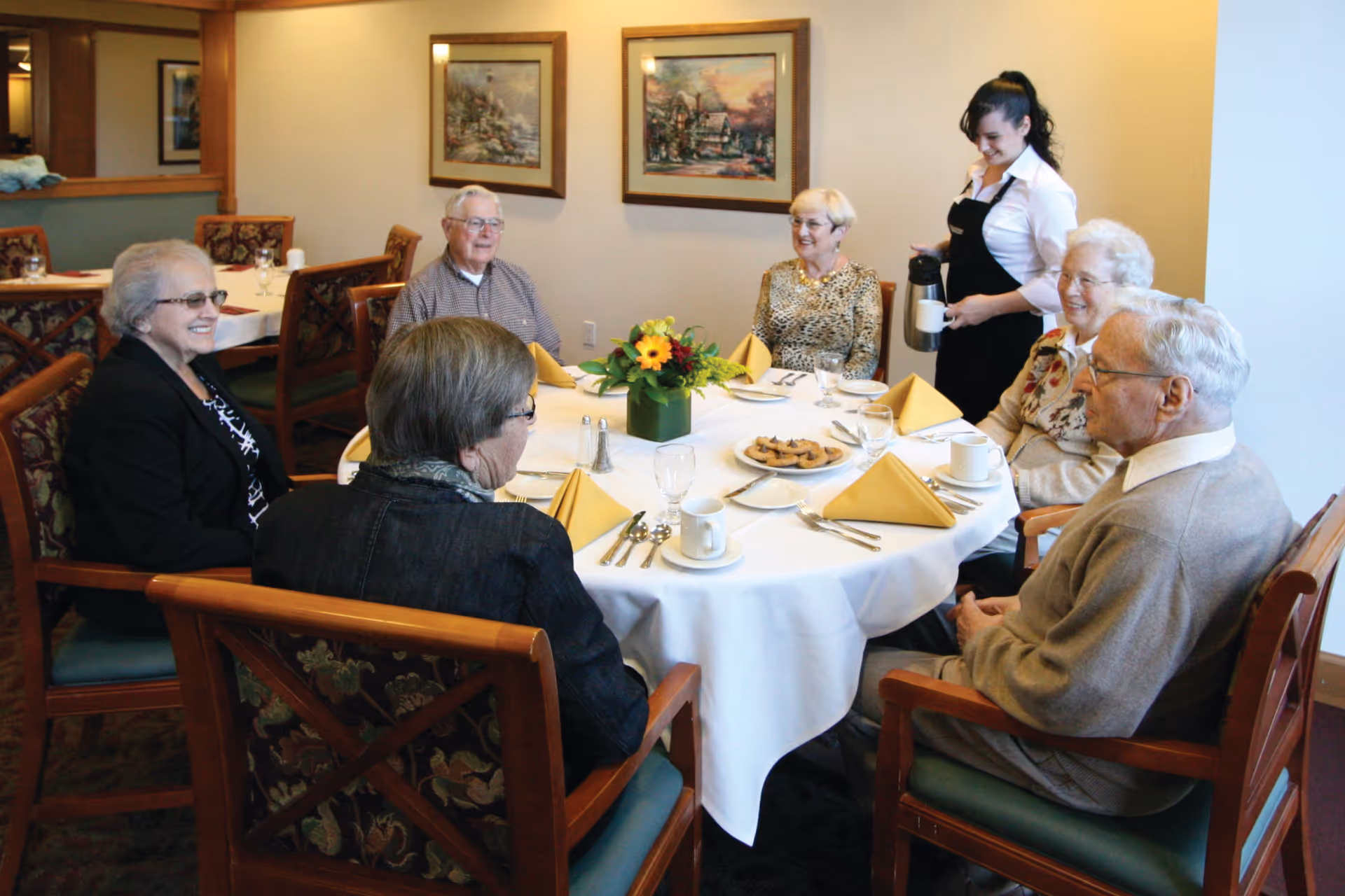 A group of six elderly people sitting around a round dining table with a white tablecloth, yellow folded napkins, cups, and a plate of cookies. A waitress is pouring coffee for one of the elderly women. The room has framed paintings on the wall and wooden chairs with floral upholstery.