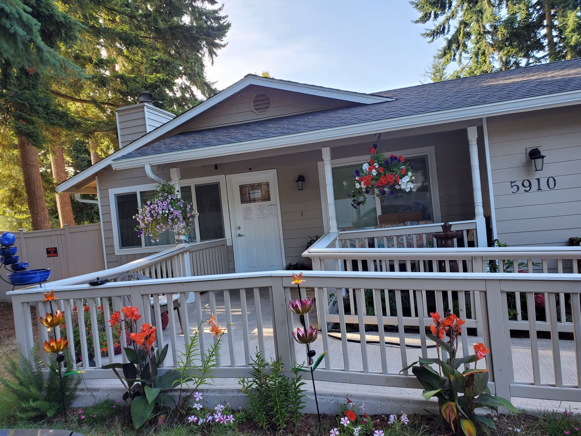 Exterior view of a single-story building with a porch and ramp entrance. The building is light gray with white trim and has the number 5910 displayed on the wall. There are hanging flower baskets on the porch and colorful flowers planted along the ramp railing. Tall trees are visible in the background.