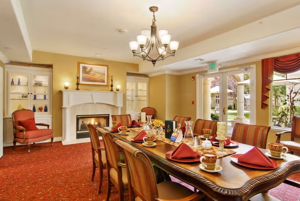 A warmly lit dining room with a long wooden table set for eight people with red napkins, cups, and plates. The room features a red patterned carpet, a white fireplace with a painting above it, two glass cabinets with decorative items, a chandelier, and large windows and doors that open to an outdoor garden area.