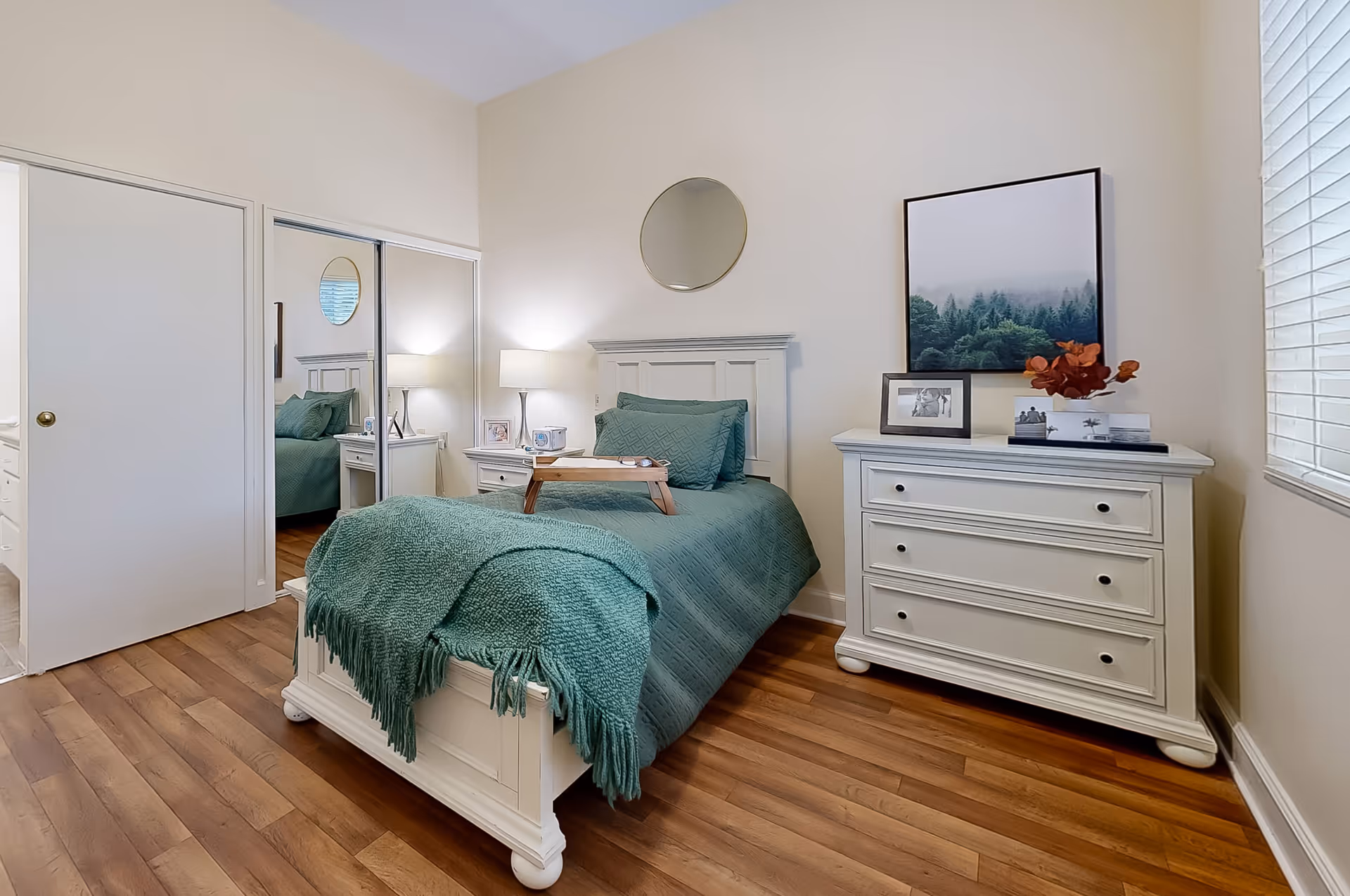 A cozy bedroom with a single white wooden bed covered with teal bedding and a matching throw blanket. A small wooden tray is placed on the bed. Next to the bed is a white nightstand with a lamp and picture frames. A white dresser with three drawers holds framed photos, a decorative plant, and a large framed picture of a misty forest. The room has wooden flooring, a round mirror above the bed's headboard, and a window with blinds on the right side. A closet with mirrored sliding doors is visible on the left.