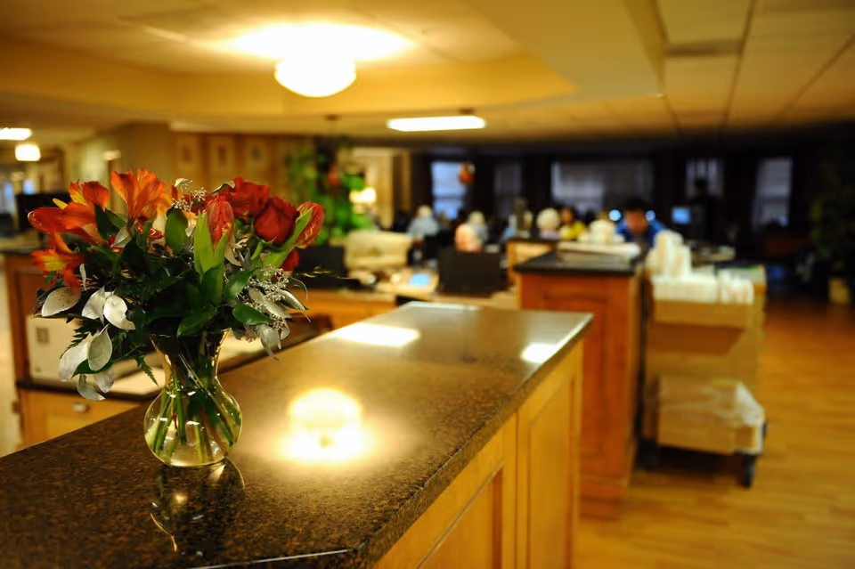 A vase with a bouquet of red and orange flowers placed on a polished countertop in a warmly lit interior space. In the background, there are people sitting and engaging in activities in a common area with wooden flooring and soft lighting.