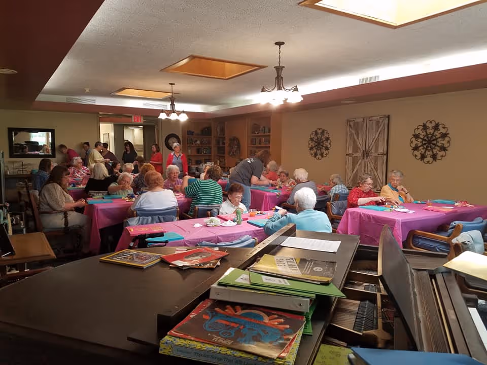 A group of elderly people seated at tables covered with pink tablecloths in a well-lit room, engaging in activities and socializing. The room has beige walls decorated with metal wall art and a wooden shutter. In the foreground, there is a piano with various books and papers on top. Several staff members are assisting the elderly.