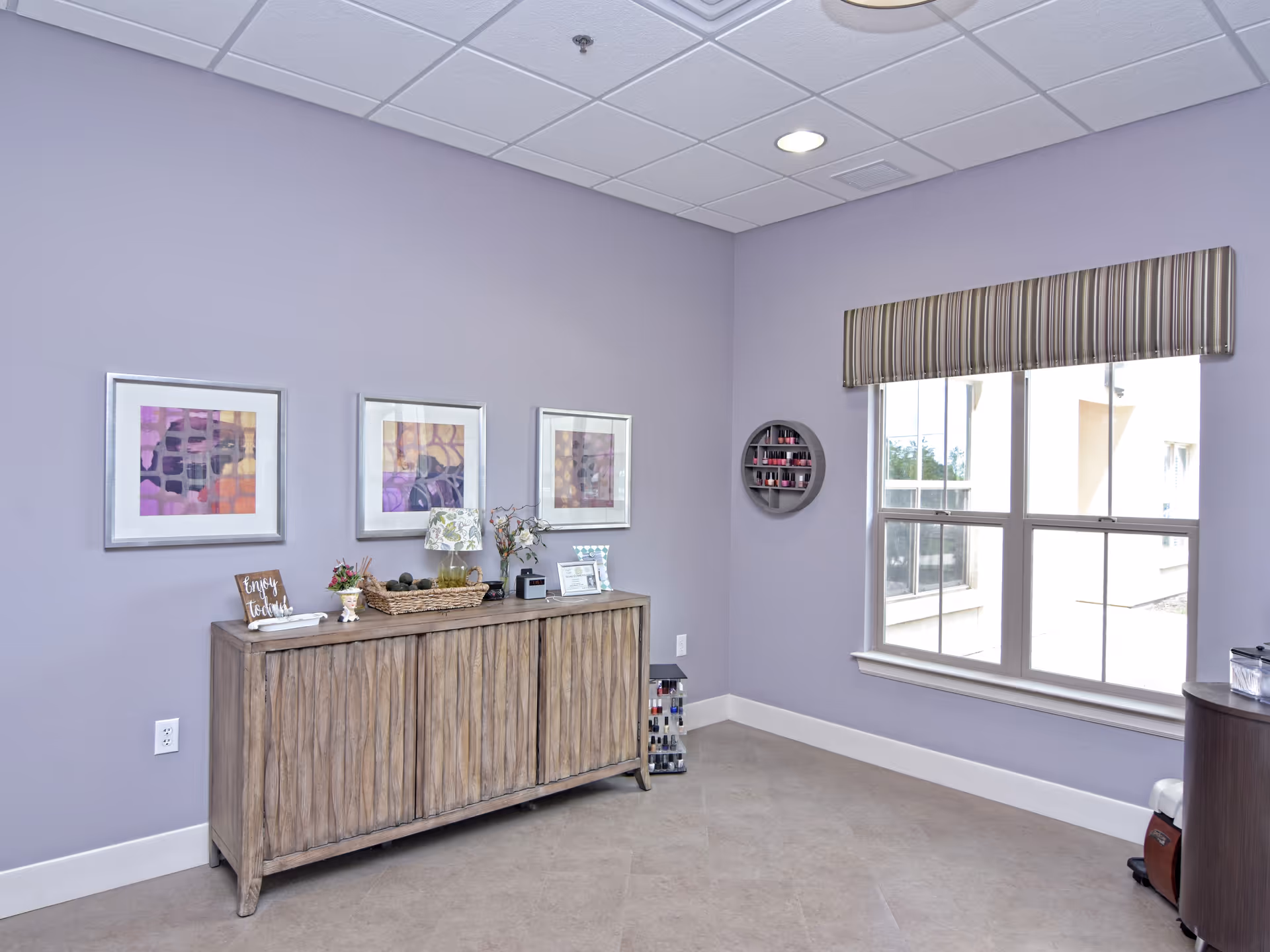 A softly lit room with lavender walls featuring a wooden sideboard against one wall. On the sideboard are decorative items including a small lamp, a basket, flowers, and framed pictures. Above the sideboard are three framed abstract art pieces with purple and orange tones. To the right, there is a large window with a striped valance, allowing natural light to enter. A circular wall-mounted shelf holds various nail polish bottles, and additional nail polish bottles are displayed on a stand on the floor near the window. The floor is tiled, and the ceiling has recessed lighting.