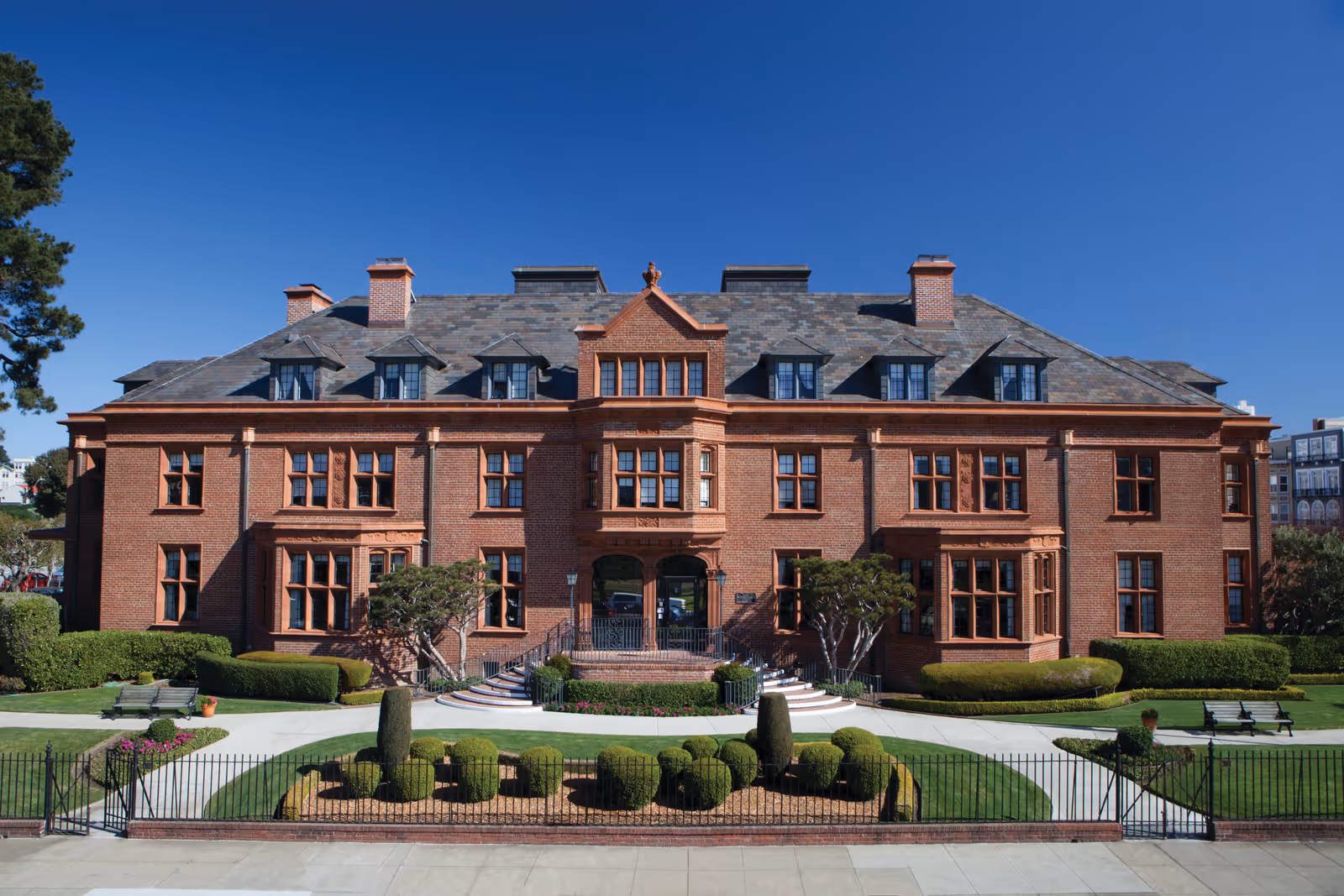 Front exterior of a large historic red-brick mansion with symmetrical windows and manicured lawns under a clear blue sky.