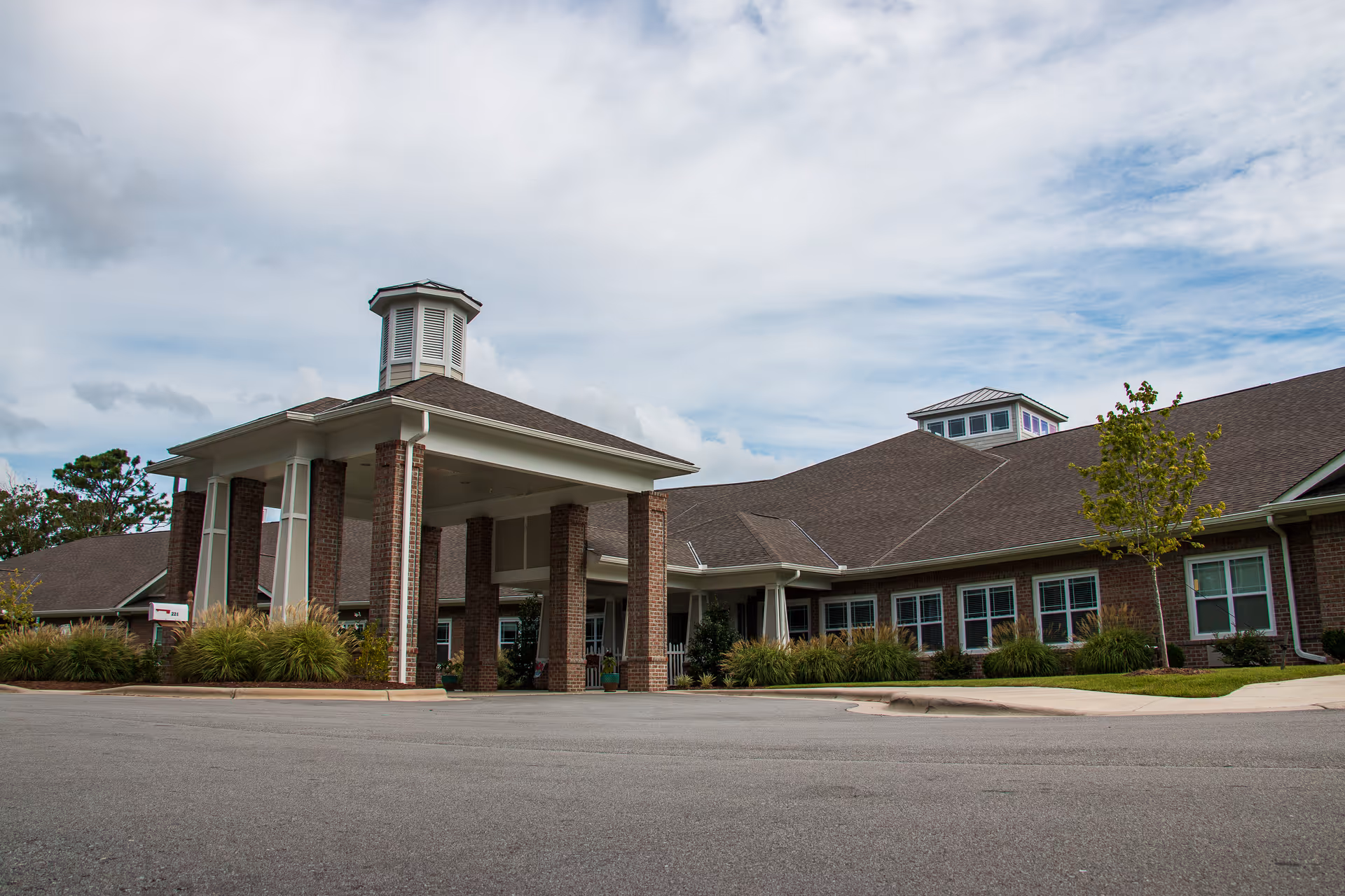 Exterior view of a single-story brick building with a covered entrance supported by brick columns, surrounded by landscaping and a paved driveway under a partly cloudy sky.