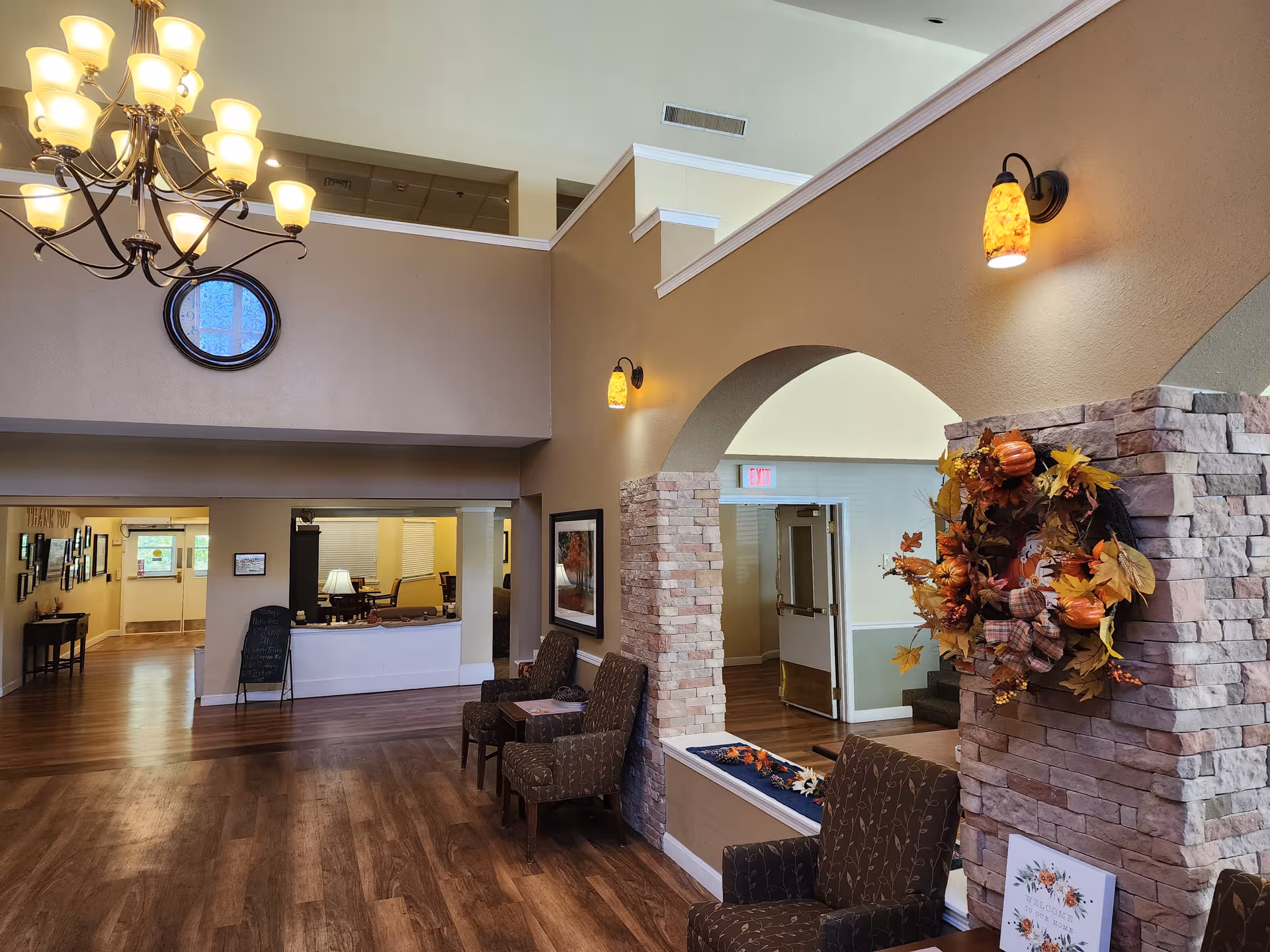 Interior view of a senior living facility lobby area with wooden flooring, beige walls, and a high ceiling. There are several cushioned chairs arranged along the walls, a decorative autumn wreath on a stone pillar, and warm wall sconces. A chandelier with multiple lights hangs from the ceiling. In the background, there is a reception desk and a hallway leading to other rooms.