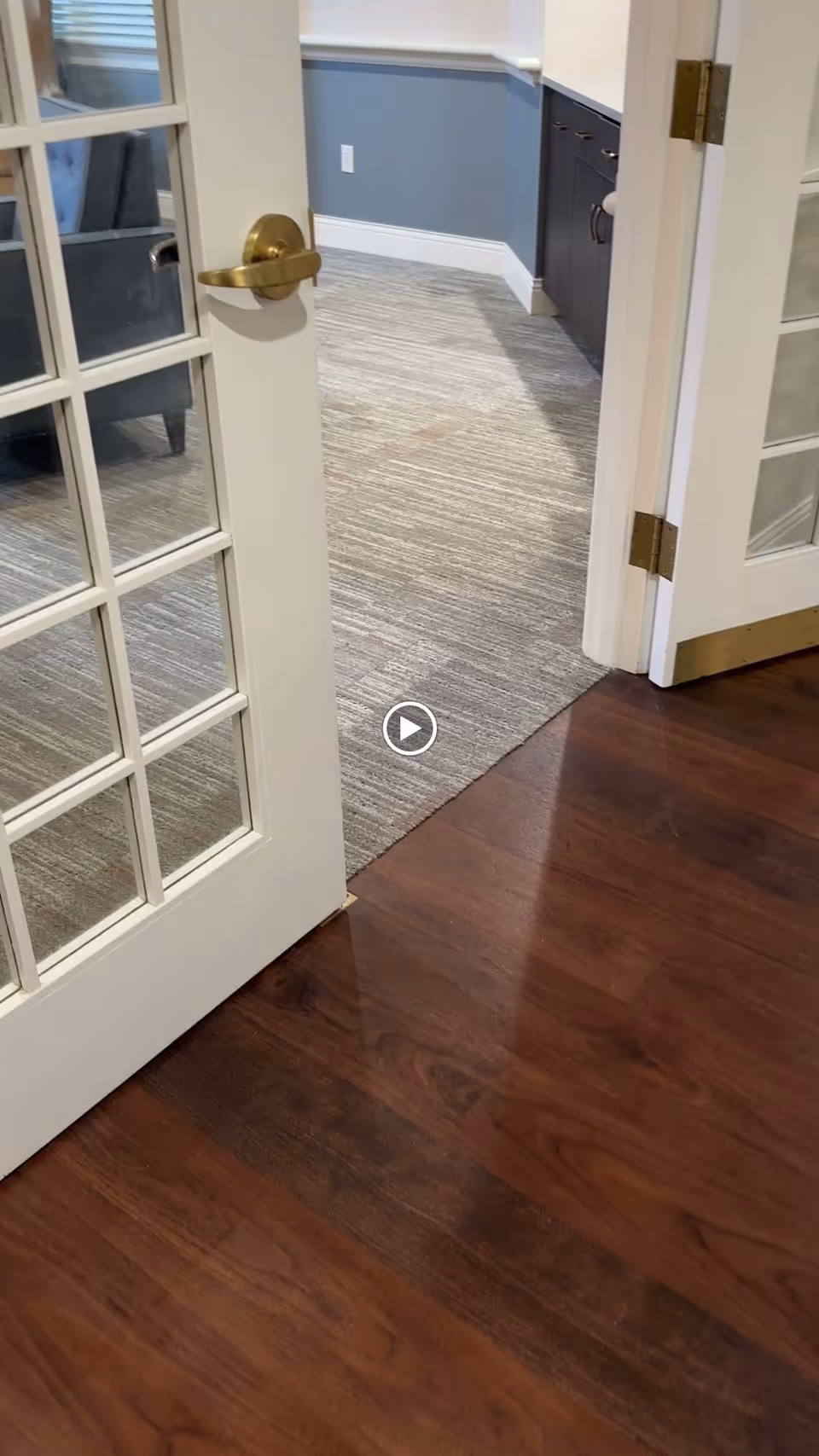 View through an open white door with glass panes into a room with gray carpet and blue-gray walls. The room has a dark wood cabinet with silver handles and a white electrical outlet on the wall. The floor outside the door is dark wood.