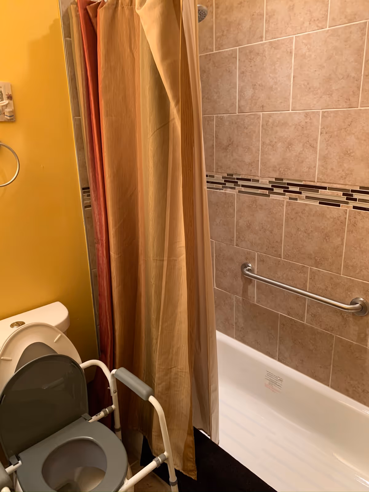 A bathroom with a toilet equipped with a raised seat and armrests for accessibility, a shower with beige tiled walls and a horizontal decorative tile strip, a metal grab bar on the shower wall, and a multi-colored shower curtain.
