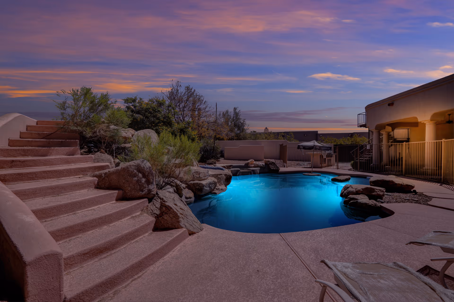 Outdoor swimming pool area at dusk with illuminated blue water, surrounded by rocks and desert plants. There are pinkish stairs on the left leading up, and a building with a covered patio and columns on the right. Lounge chairs are visible in the foreground.