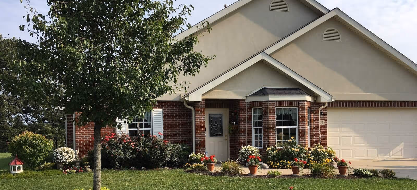 Front exterior view of a single-story house with a brick and beige facade, a white front door with decorative glass, a two-car garage, and a well-maintained garden with various flowering plants and a tree in the front yard.