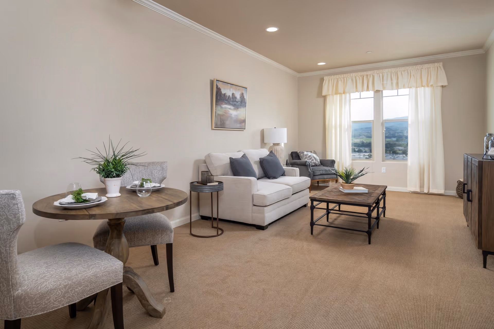 A cozy living room with beige walls and carpet, featuring a small round wooden dining table set for two with plants and glassware, a white sofa with gray cushions, a side table with a lamp, a gray armchair, a wooden coffee table with a plant and book, a wooden cabinet, and large windows with cream curtains letting in natural light.