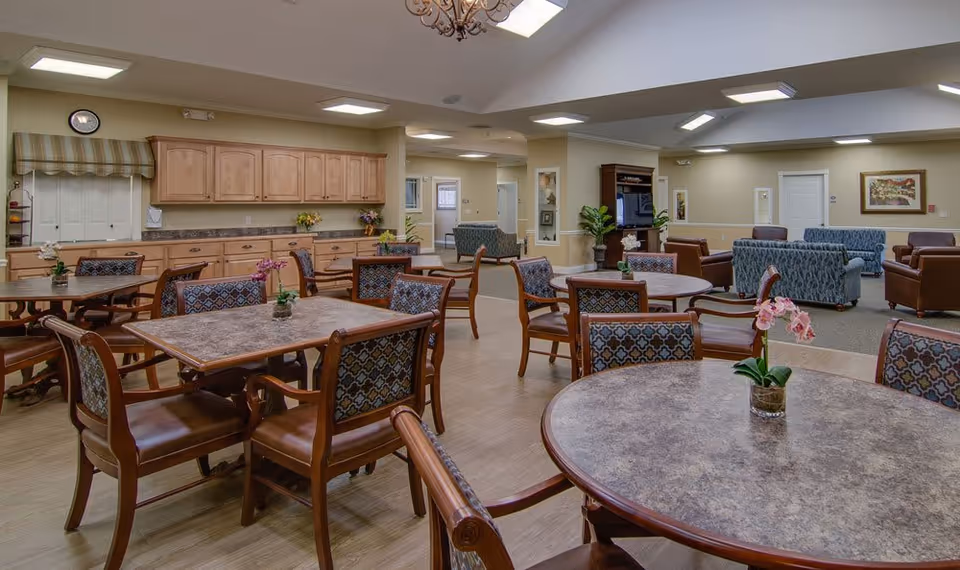 A spacious senior living facility common area with multiple round and rectangular tables surrounded by wooden chairs with patterned cushions. The room features a kitchenette with wooden cabinets and a countertop along one wall. In the background, there is a seating area with sofas and armchairs arranged around a TV. The ceiling has recessed lighting and skylights, and the walls are decorated with framed artwork and plants.