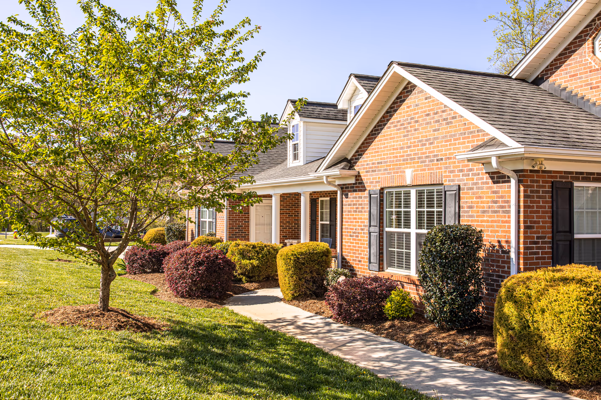 Exterior view of a brick residential building with black shutters and a gray shingled roof, surrounded by neatly trimmed bushes and a tree on a sunny day with a clear blue sky.