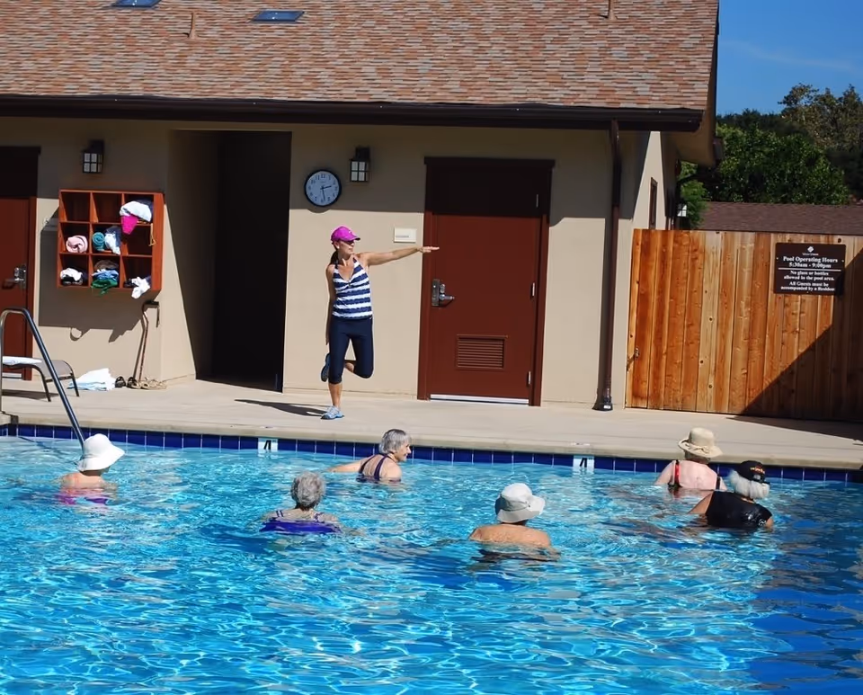 A group of elderly people participating in a water exercise class in an outdoor swimming pool. An instructor stands on the pool deck demonstrating a stretch, wearing a pink cap and striped tank top. Behind them is a building with a clock and a wooden fence with a sign displaying pool operating hours.