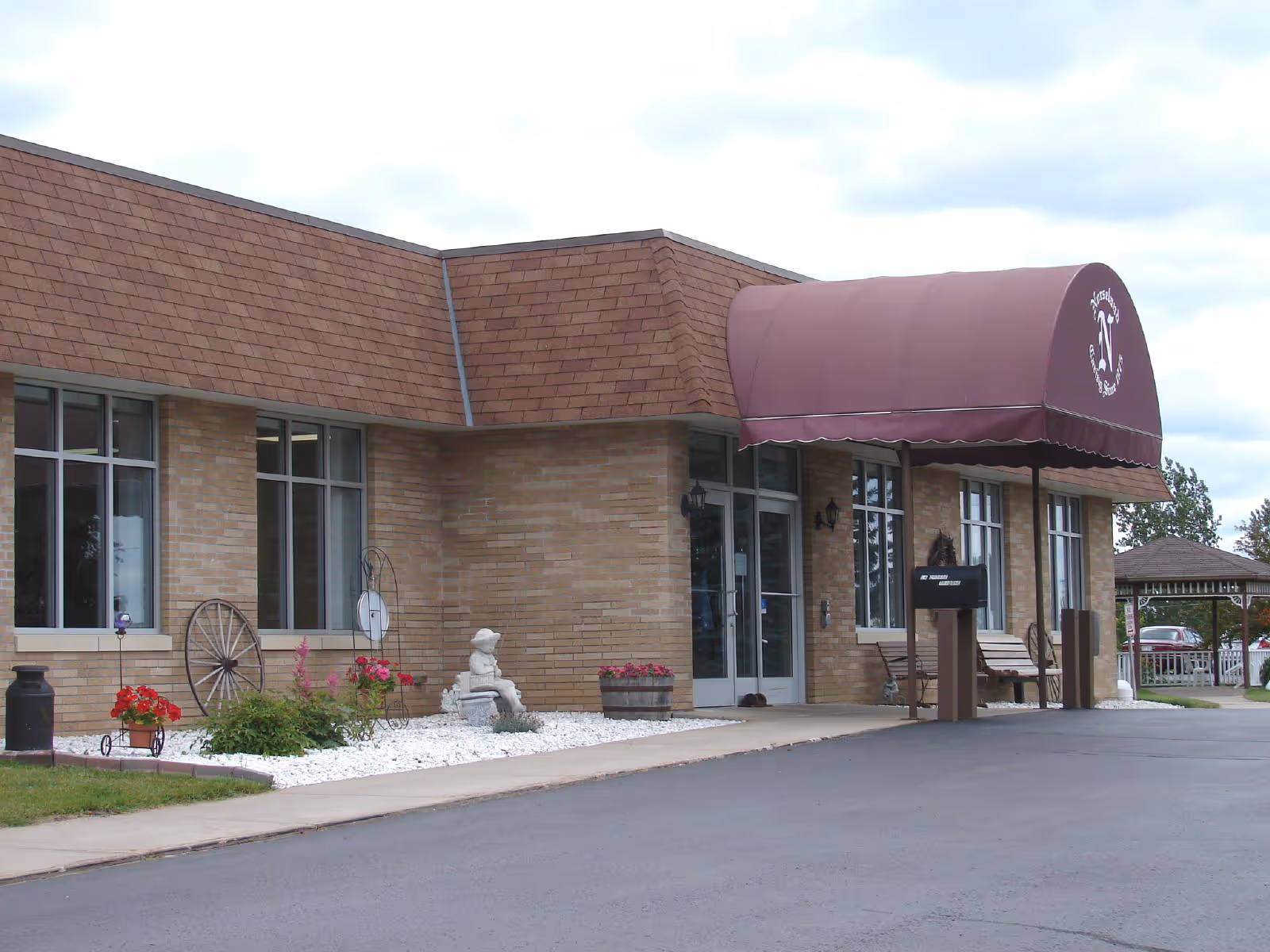 Exterior view of Norseland Nursing Home showing a brick building with large windows, a maroon awning over the entrance, a small garden with flowers and decorative items, and a paved driveway.
