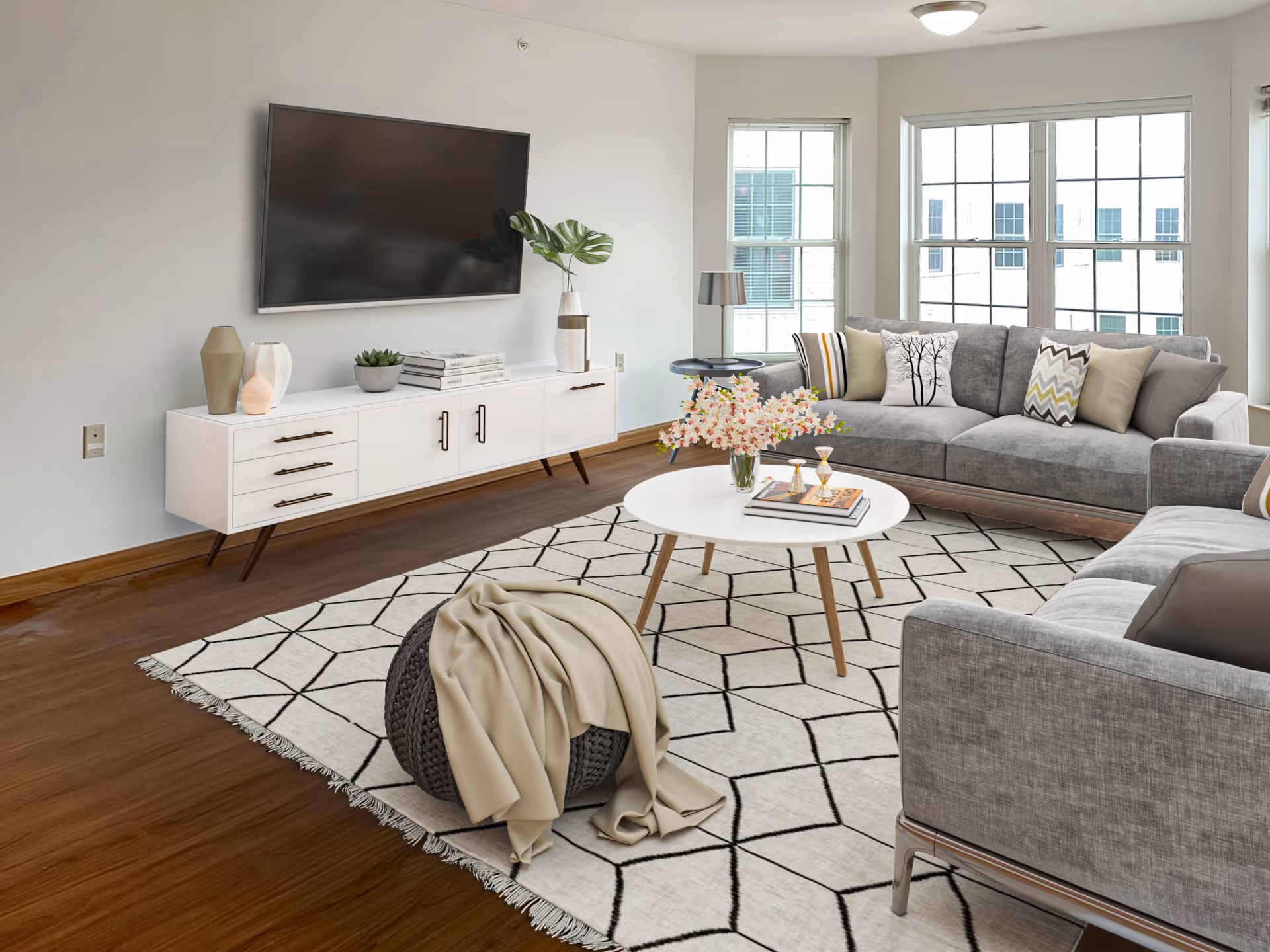 Bright living room with gray sofas, a wall-mounted TV, white media console, round coffee table on a patterned rug, and large windows.