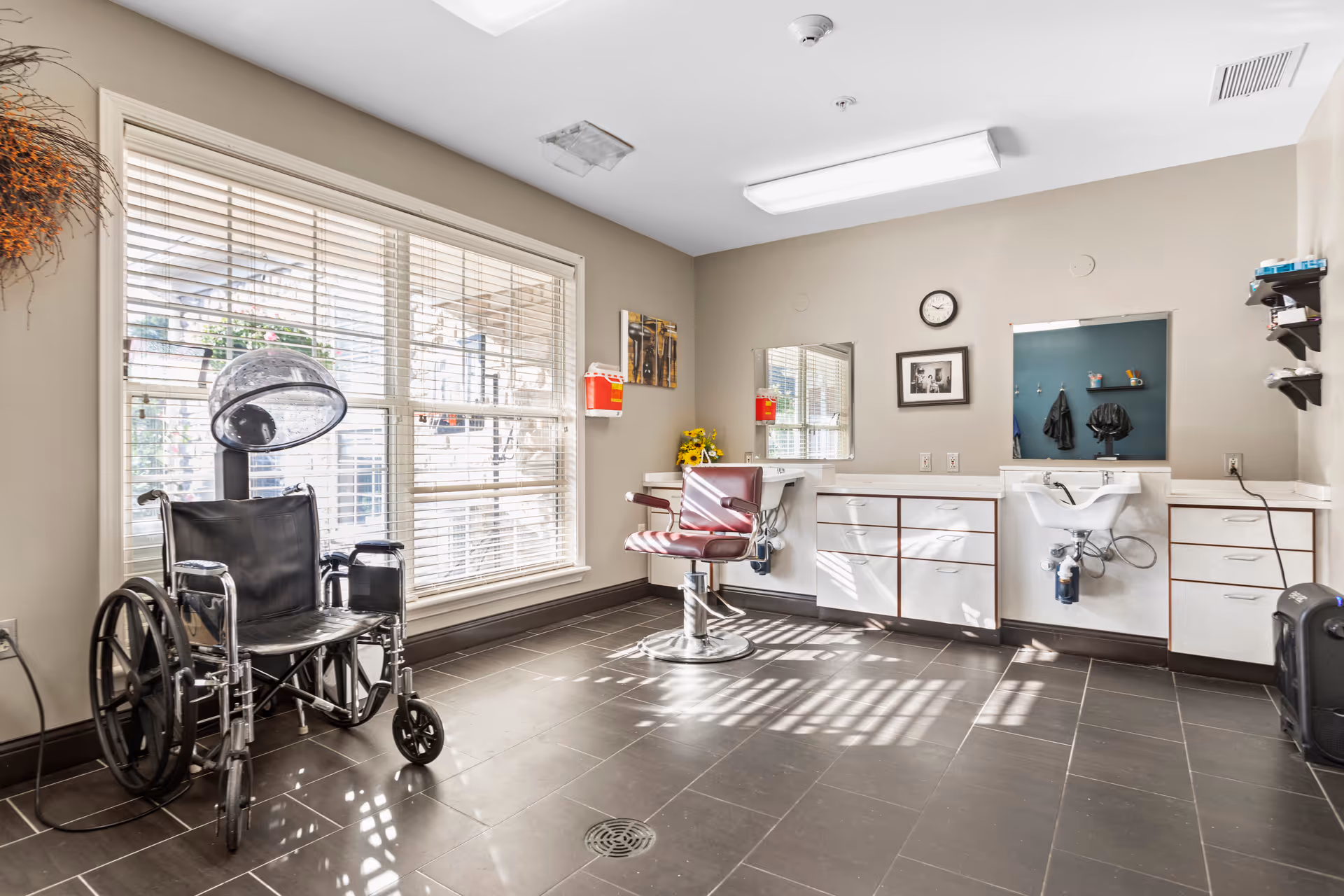 A bright room with large windows covered by blinds, featuring a wheelchair and a hair drying station with a hood dryer. There is a salon chair in front of a counter with drawers and a mirror, and a small sink with plumbing visible underneath. The walls are beige, and the floor is tiled in dark gray. A clock and a framed picture hang on the wall above the counter.