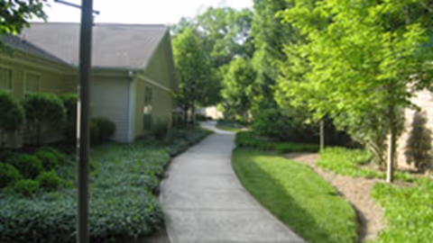A paved walkway winding through a landscaped garden area with green grass, bushes, and trees on both sides, adjacent to residential-style buildings.