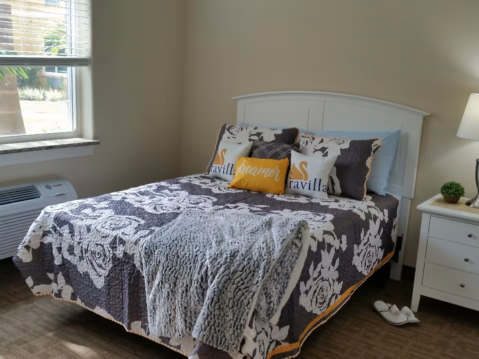 A neatly made bed with a white headboard in a bedroom. The bed has a dark floral-patterned quilt with white flowers and several decorative pillows, including two with the word 'Aravilla' and one yellow pillow with the word 'dreamer'. A gray textured blanket is draped over the foot of the bed. Next to the bed is a white nightstand with a small green plant and a lamp. A window with blinds is on the left side, and a pair of white slippers is on the floor near the nightstand.