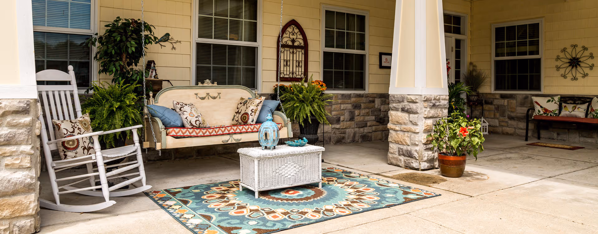 Covered front porch with a rocking chair, hanging loveseat, wicker coffee table on a colorful rug and potted plants.