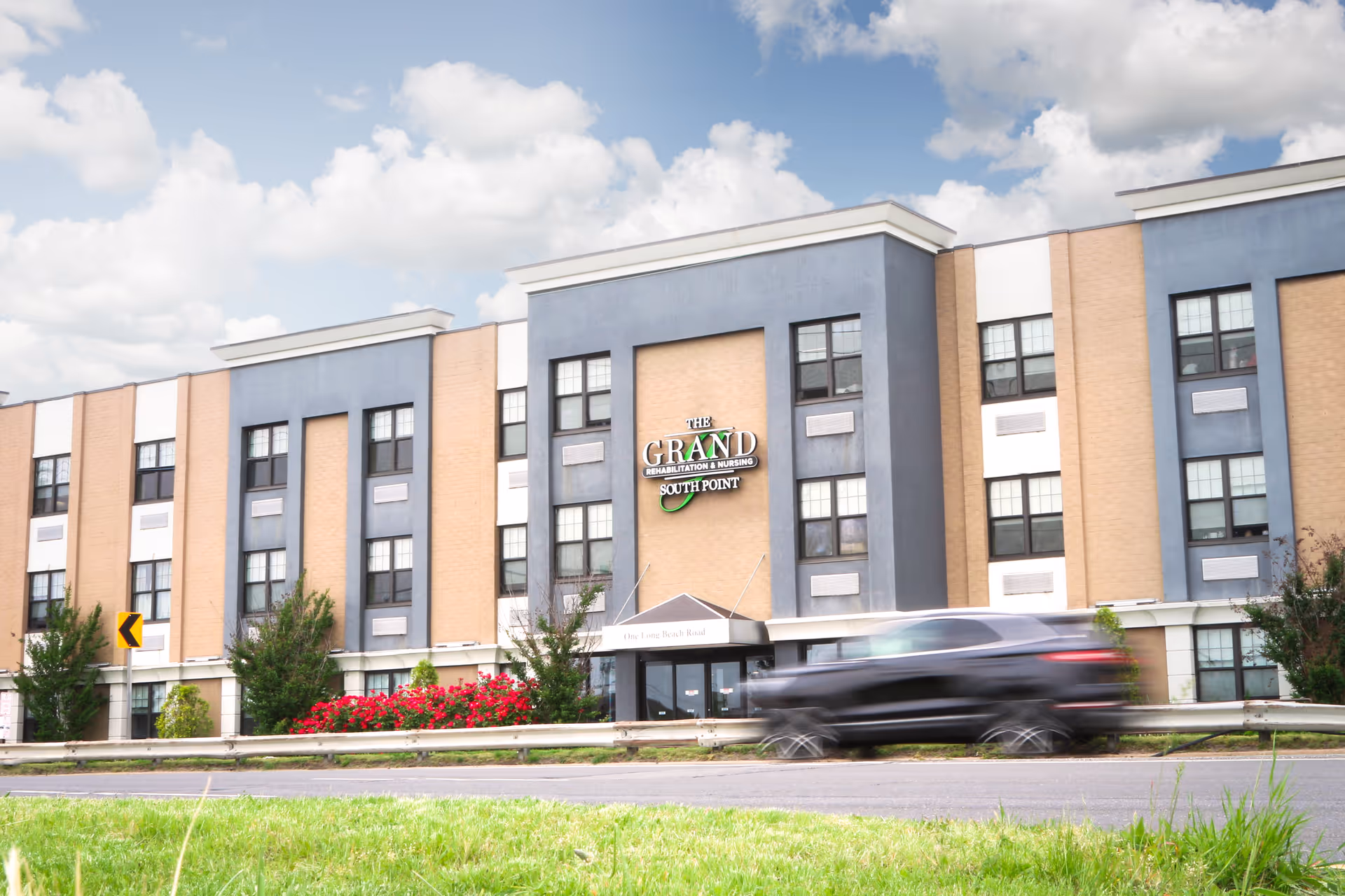 Exterior view of The Grand Rehabilitation and Nursing at South Point building with a multi-story facade featuring beige and gray panels, multiple windows, and a sign displaying the facility's name. A black SUV is driving past on the road in front of the building, with green grass and red flowers visible in the foreground under a partly cloudy sky.