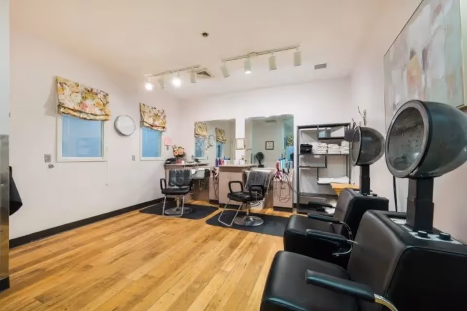 Interior view of a hair salon area in a senior living facility with wooden floors, two salon chairs in front of mirrors, hair dryers with black chairs, a shelf with towels, and floral window valances.