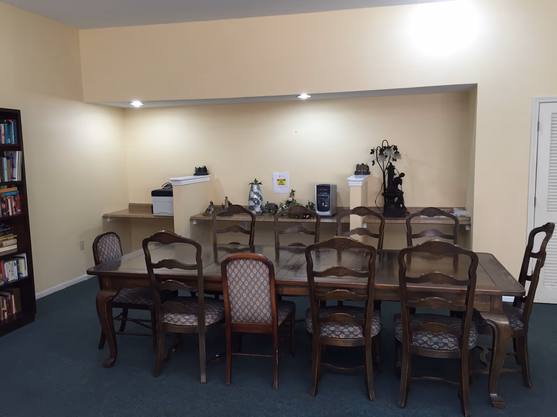 A room with a large wooden table surrounded by ten chairs with patterned cushions. The walls are painted light yellow, and there is a built-in counter along the back wall with decorative items, a printer, and a computer tower. A bookshelf filled with books is visible on the left side, and a closed white door is on the right.