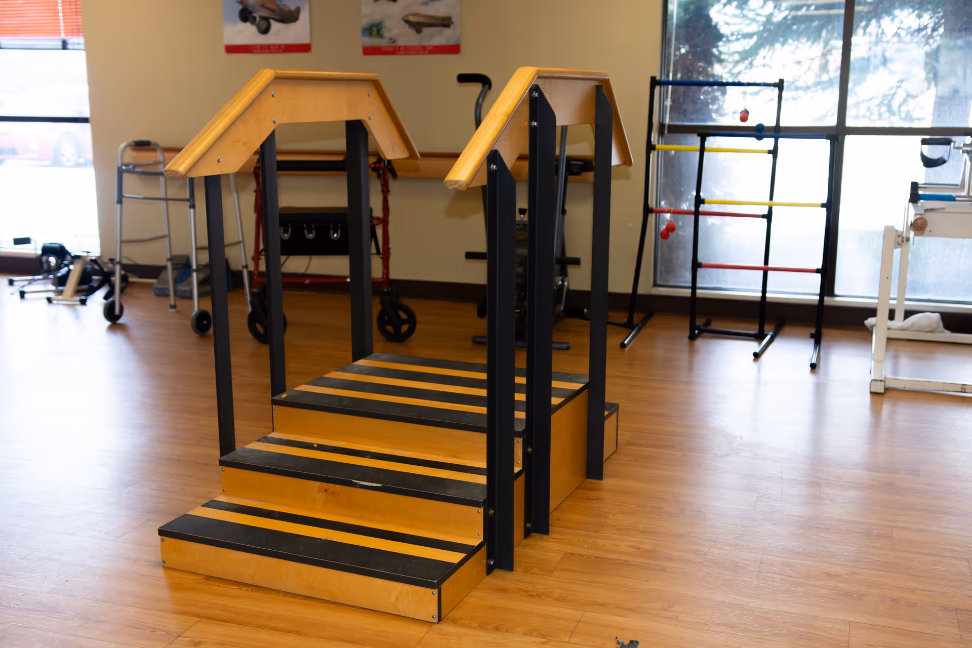 Indoor physical therapy room with wooden stairs equipped with handrails for rehabilitation exercises. In the background, there are walkers, exercise equipment, and large windows letting in natural light.