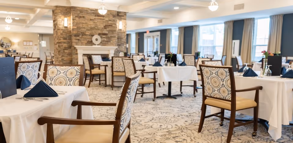 Bright, spacious dining room with neatly set tables, patterned chairs, and a stone fireplace.