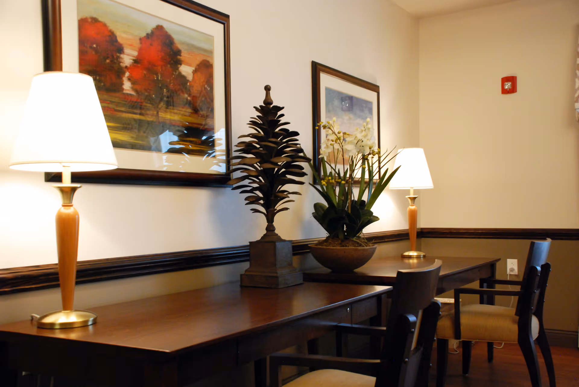 A well-lit interior seating area with a wooden table, two lamps, framed artwork, decorative plants, and chairs.