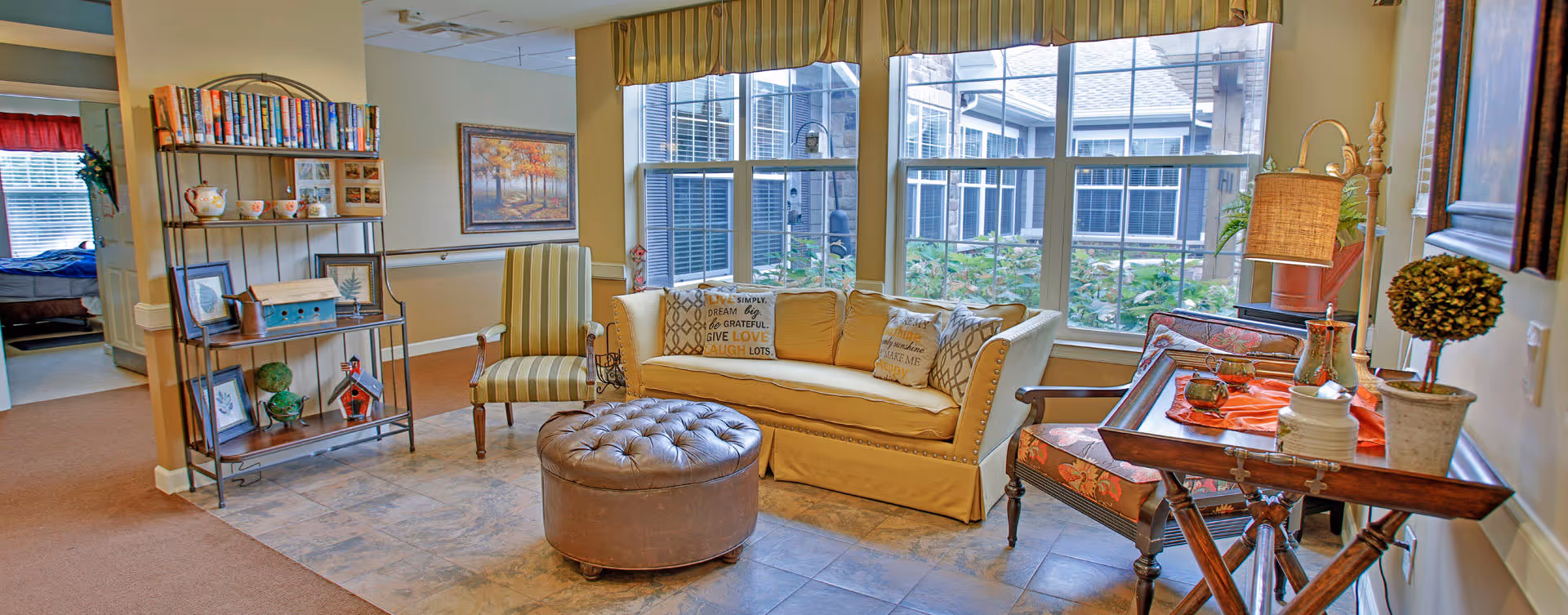 A cozy living room area with a beige sofa adorned with decorative pillows, a striped armchair, and a round brown ottoman. Large windows with yellow striped valances let in natural light. A wooden side table with a lamp, potted plant, and decorative items is on the right. A metal shelving unit with books and decorative pieces is on the left. In the background, a bedroom is partially visible through an open doorway.