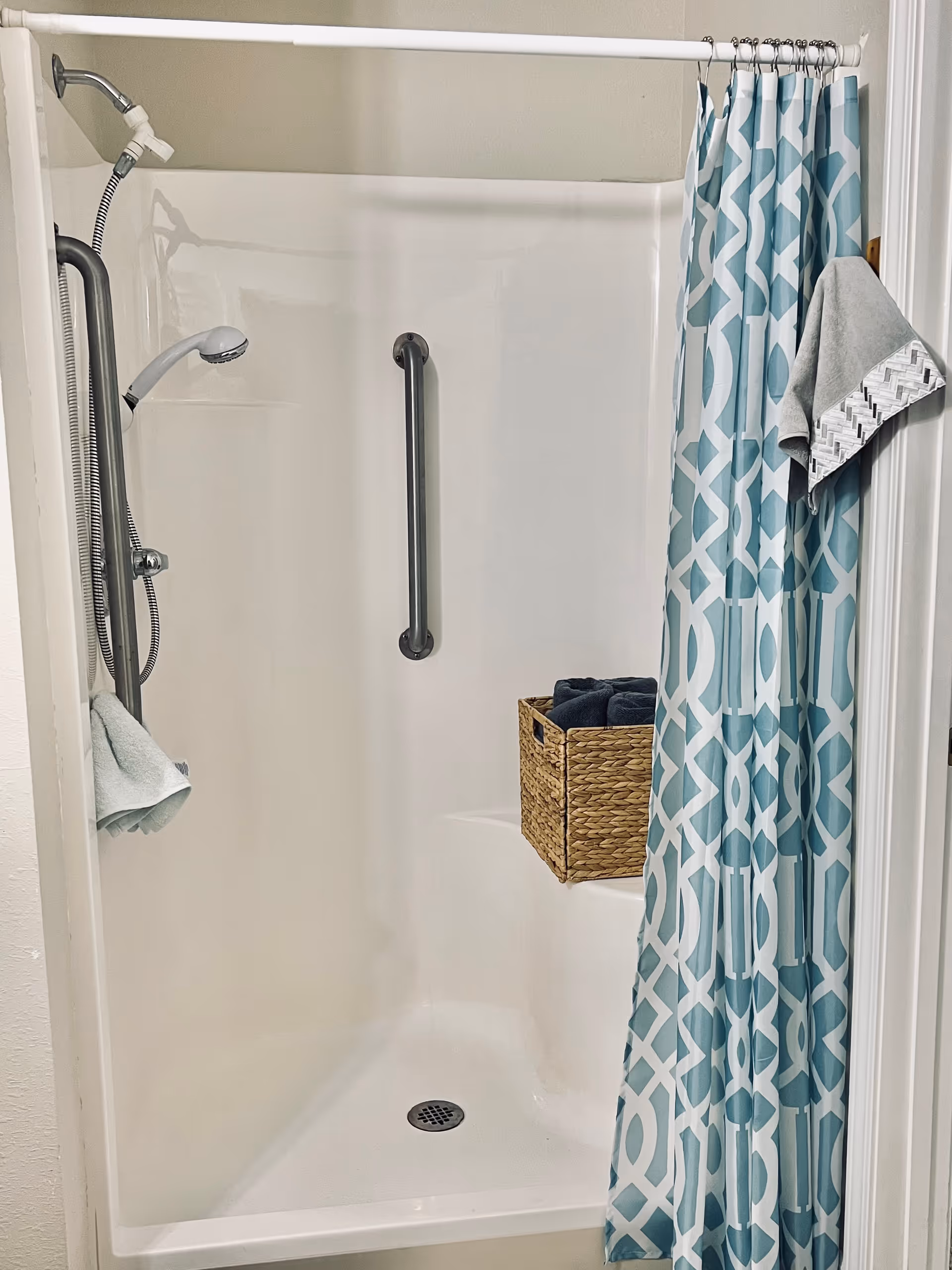 A white shower stall with a blue and white patterned shower curtain partially drawn to the side. Inside the shower, there is a silver grab bar on the wall, a handheld showerhead attached to a flexible hose, and a built-in seat holding a woven basket with rolled dark towels. A gray towel is hanging on a hook outside the shower.