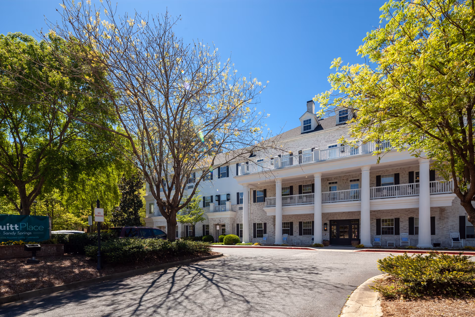 Exterior view of PruittPlace - Sandy Springs facility showing a large white building with multiple windows, black shutters, and a covered porch supported by white columns. Trees with green and some bare branches surround the driveway leading to the entrance under a clear blue sky.