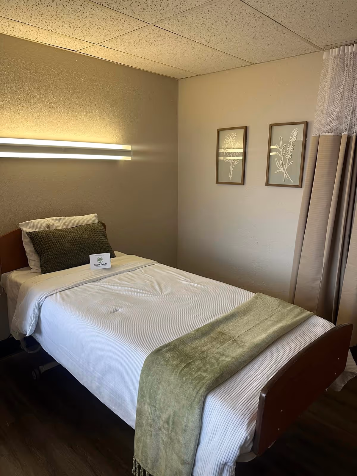 A single bed in a room with white bedding, a green throw blanket at the foot, and a green pillow. Above the bed is a modern horizontal light fixture. Two framed botanical prints hang on the beige wall to the right. A beige curtain is partially visible on the right side of the image. A small card with the Grace Pointe Wellness Center logo is placed on the bed.